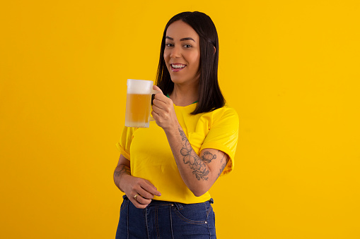Young beautiful woman in yellow color shirt from Brazil holding a glass of beer in studio shot with various expressions of feelings