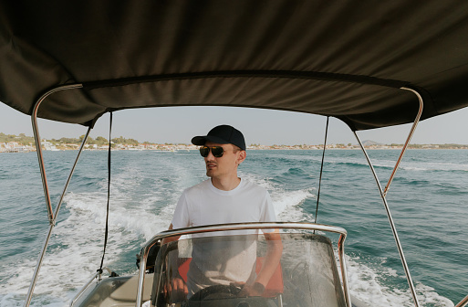 One young Caucasian guy in a cap and sunglasses sails alone on the sea on a boat, steering the helm and looking to the left, on a sunny summer day, close-up side view.