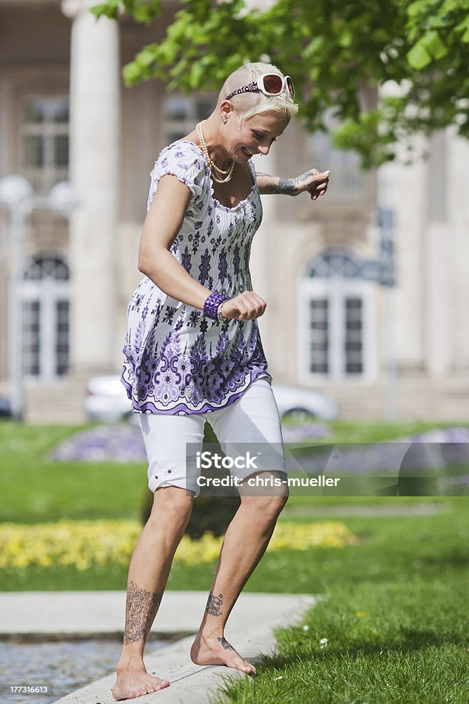 young woman at a pond in the park pretty young woman with various tattoos stands bare feet on the banks of a pond in the park Adult Stock Photo