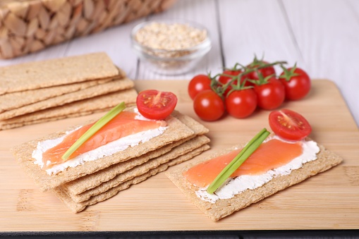 Tasty crispbreads with salmon, cream cheese, green onion and tomato on white wooden table, closeup