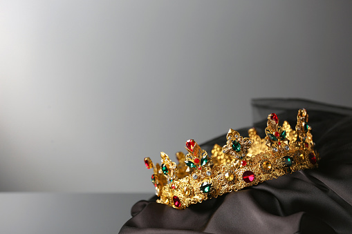 Beautiful golden crown with gems on dark cloth against grey background. Space for text