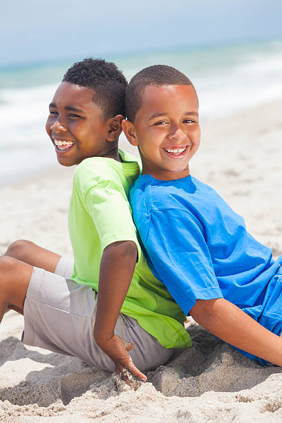 Two young African American boys sitting back to back playing having fun on a sunny beach