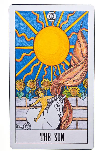 The sun tarot card isolated on a white background.  The card's top part is taken up by a yellow sun with squiggly lines coming out of the circle center to represent the rays of a sun.  The sun is set on a light blue sky.  A row of sunflowers is sitting on a stone wall beneath the sun.  A yellow naked child is sitting on a white horse in front of the stone wall.  The child's arms and legs are extended.  A brownish pink flag is flying from behind the horse.