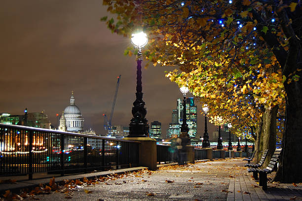 Riverside footpath leading away from camera and lit by street lamps.  London skyline including St Paul's cathedral can be seen in the background.