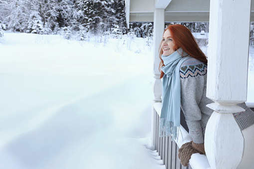 Beautiful young woman looking out from wooden gazebo on snowy day outdoors, space for text. Winter vacation