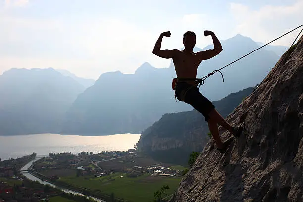 "Silhouette of a rock climber flexing biceps against picturesque view of Lake Garda, Arco, northern Italy"