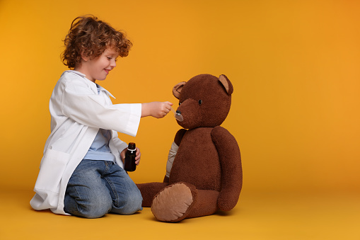 Little boy playing doctor with toy bear on yellow background. Space for text