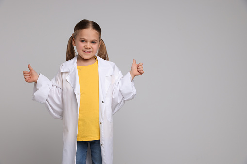 Little girl in medical uniform showing thumbs up on light grey background. Space for text