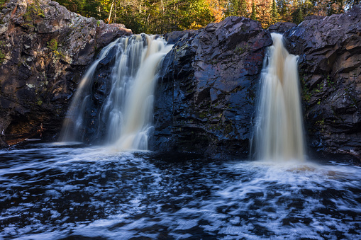 A scenic landscape featuring a twin waterfall in the woods during autumn.