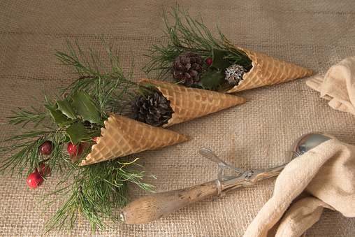 Diagonal row of waffle cones stuffed with festive greenery, berries, and pinecones on a burlap cloth.
