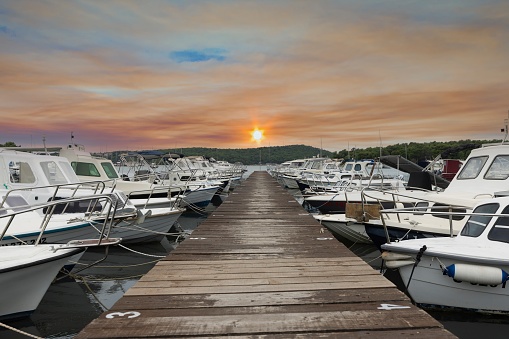 View along a wooden jetty in a harbor with docked ships in front of the setting sun in summer