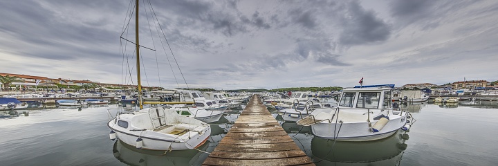 View along a wooden jetty in a harbor with docked ships in front of clouded sky in summer