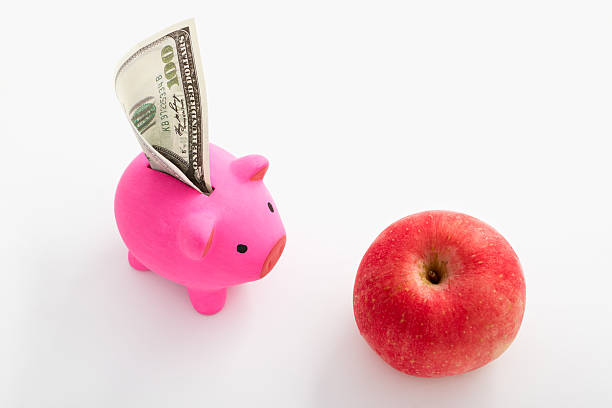 Pink piggy bank with dollar facing an red apple over white background