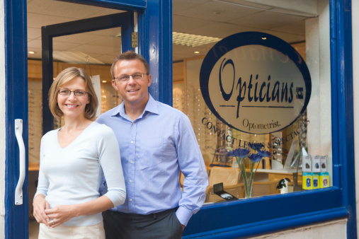 Couple standing at front entrance of optometrists wearing glasses smiling