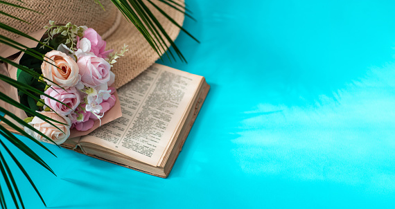 Palm leaf, pink roses, open book, straw hat are set on pastel blue background. Abstract, elegant summer composition. Relaxing vacation concept. Mood for vacation and travel. Artwork mockup with copy space