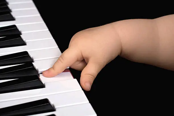 A small child play with one Finger on a Piano in front of a dark Background.You can see the forefinger and the thumb and the underarm. She is hitting one white piano key.