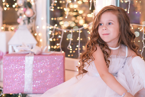 Little beautiful girl sitting on the windowsill, looking and smiling on lights background. New Year. Christmas. High quality photo