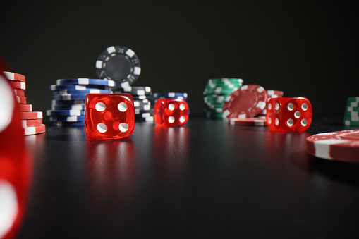 Red dice with digits and colorful poker chips scattered on dark table leaving reflections. Equipment for playing professional gambling games