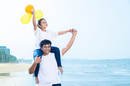 Happy indian father carrying his daughter with balloon in hands on shoulders in front of the beach enjoying holiday, vacation.