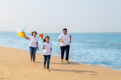 Happy indian Family of three enjoying summer vacation. Man and woman with their daughter running with balloon and paper windmill on beach.