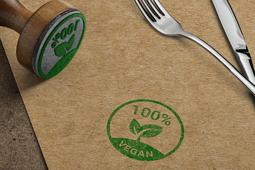 A close-up shot of a 100% vegan stamp on an eco-friendly brown napkin, placed alongside a polished silver fork and knife with a modern handle, symbolizing sustainable and ethical dining choices.