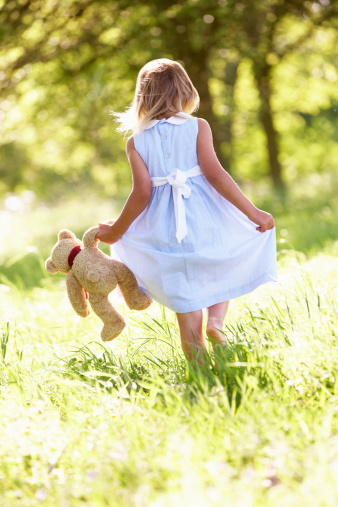 Back View Of Young Girl Walking Through Summer Field Carrying Teddy Bear