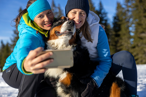 Two carefree women friends taking selfie using mobile phone camera with Bernese mountain pet dog on snowy slope