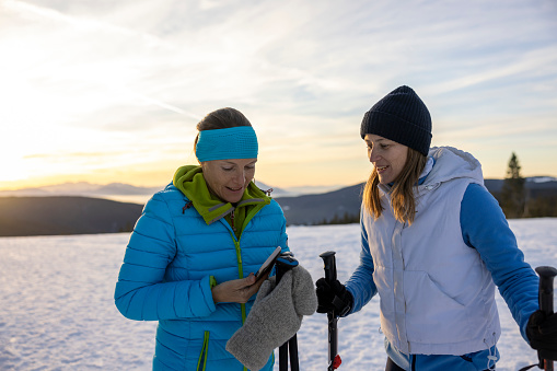Two women friends in warm clothing holding ski poles and using mobile phone on snowy winter landscape