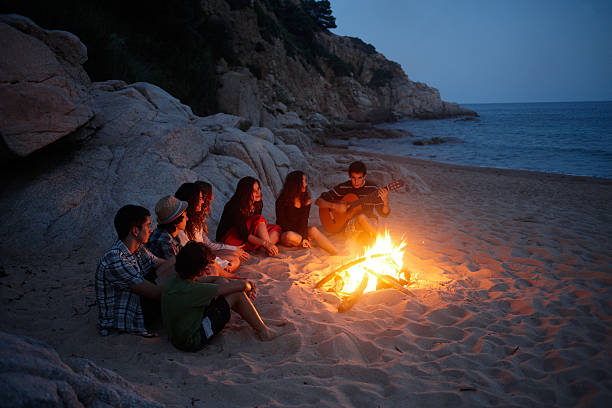 adolescenti cantare attorno a un fuoco in spiaggia - friendly fire foto e immagini stock