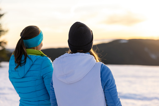 Rear view of two women in warm clothing admiring mountain view during ski holiday from snowy winter landscape