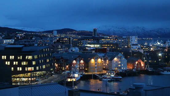 A night view of a tromso city on a harbor with snow-capped mountains in the background. The lights from the buildings and boats create a contrast with the dark sky and water.