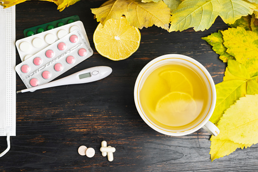 Tea cup with lemon, thermometer, capsules, pills, medicine in a blister pack, medical mask on a wooden table with yellow leaves. Concept of treatment autumn cold, flu and coronavirus, top view.