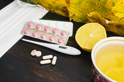 Capsules, pills, medicine in a blister pack, thermometer, tea cup with lemon on a wooden table with yellow leaves. Concept of treatment autumn cold, flu and coronavirus