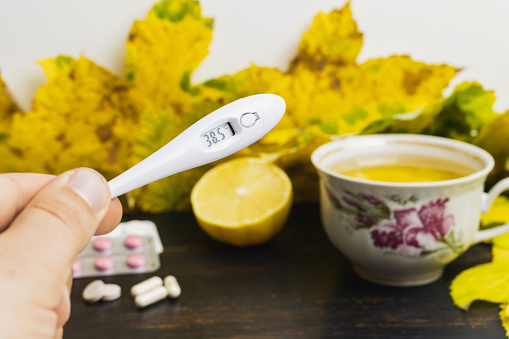 Male hand holds digital thermometer indicates high temperature 38.5, above table with tea cup, lemon, capsules, pills and yellow leaves. Concept of treatment fever, autumn cold, flu and coronavirus.