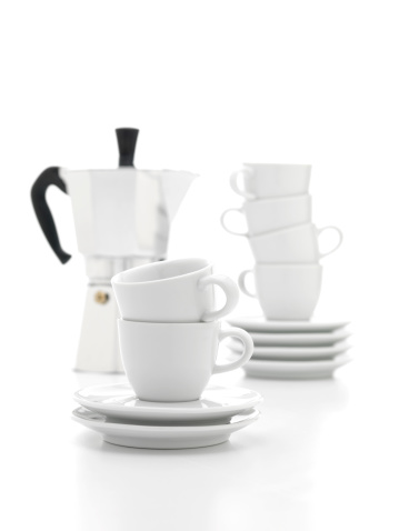 Two white cups and two white saucers stacked in front of a coffee percolator and more cups and saucers.