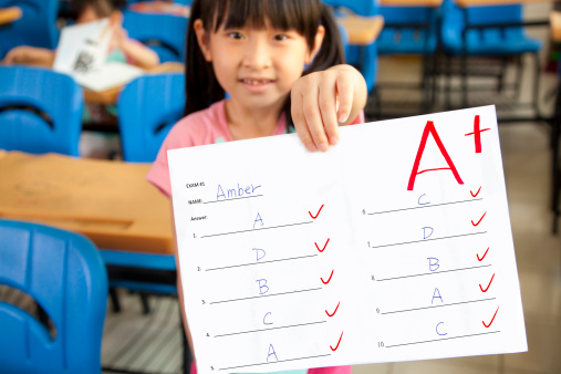 smiling asian little girl showing exam paper with a plus in the classroom