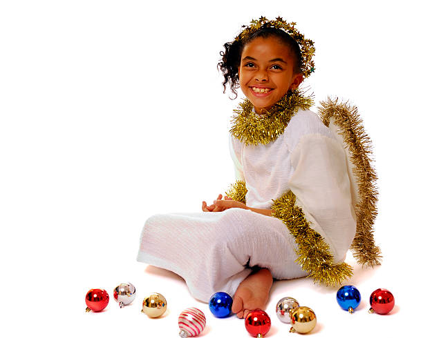 Isolated.  Smiling, young Bi-racial girl in angel costume sitting among scattered Christmas bulbs.