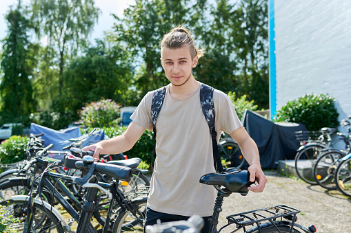 Teen guy college student with backpack with outdoor bike, educational building background, bike parking. Youth, transport, students lifestyle concept