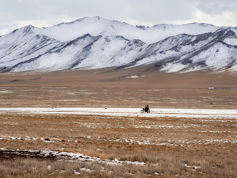 Beautiful Mongolian winter landscape snow mountain with a small figure of a ride on a black motorcycle through a wide snow-covered valley against the backdrop of high mountains.