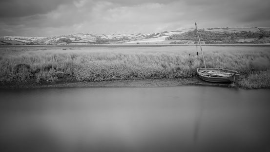 Embark on a journey into the mystic allure of Laugharne, Wales, with this captivating black and white landscape photograph, skillfully converted from an infrared long exposure. Set against the tranquil backdrop of the creek, the image unveils a solitary row boat, eerily stranded along the water's edge. The prolonged exposure imparts a surreal, almost ethereal quality, enveloping the scene in a haunting and mysterious atmosphere. This compelling composition captures the essence of both stillness and motion, evoking a sense of quiet unease. Perfect for projects seeking a blend of eerie tranquility and enigmatic allure, this photograph is a testament to the artistry of nature.