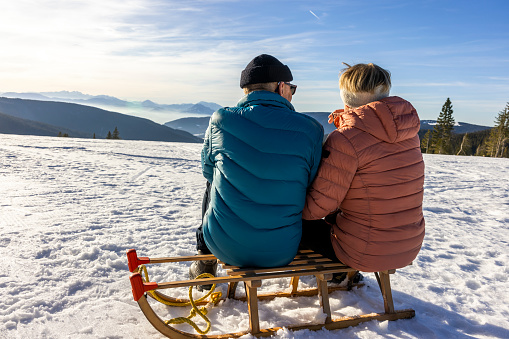 Rear view of senior couple relaxing on sled and admiring beautiful mountain view from snowy winter landscape