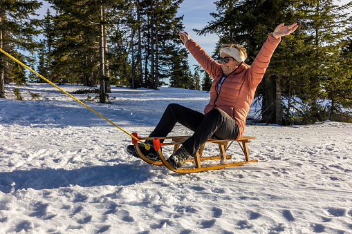 Cheerful senior woman in warm clothing having fun sledding on snowy winter hill