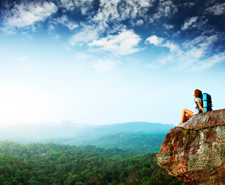 Young woman with backpack sitting on an edge of cliff and looking to a sky