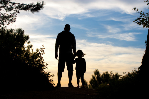 Silhouette of father and daughter holding hands watching the sunset on the top of a forest path