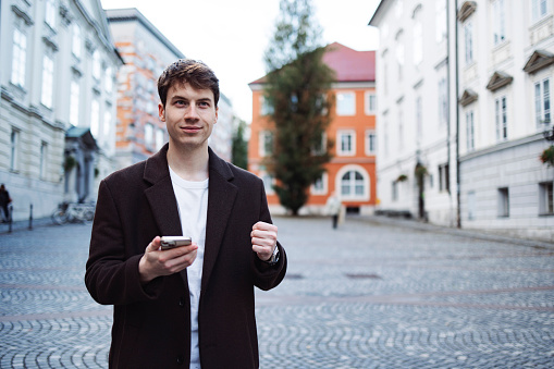 Young adult holding mobile phone in right hand and raised a fist with left hand in city center. Portrait shot of young male.