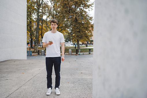 Young adult man standing on main square in city center, holding mobile phone and looking into camera. Portrait shot,