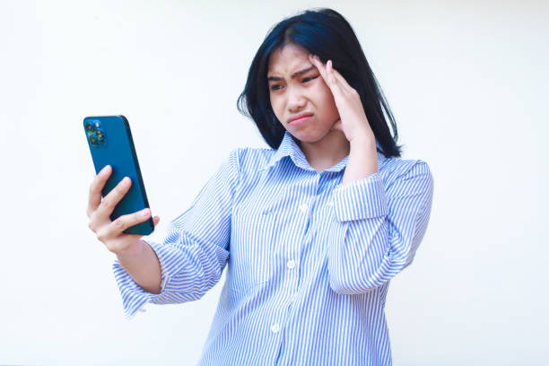 istock thoughtful asian woman using smartphone with hands on head thinking problem wearing blue striped casual shirt standing over isolated white background, look away 1774363289