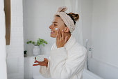 Happy young woman in bathrobe applying face cream while standing in bathroom