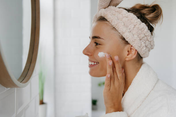 istock Attractive young woman in bathrobe applying face cream and smiling while standing in bathroom 1774418918
