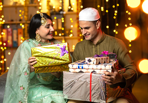 Young husband wearing skull cap giving surprise gift to excited wife in illuminated home on occasion of traditional festival